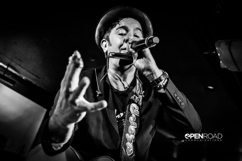 G. Love singing into a microphone he's holding while the other hand is outstretched. 