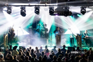 Bright lights surround Daniel Donato and Cosmic Country on the stage. Taken from the back and above so you see the crowd below and the full stage.