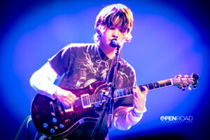 Mei Semones stands behind a microphone playing guitar and singing