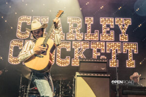 Charley Crockett raises his guitar while playing. His name emblazoned in lights behind him.