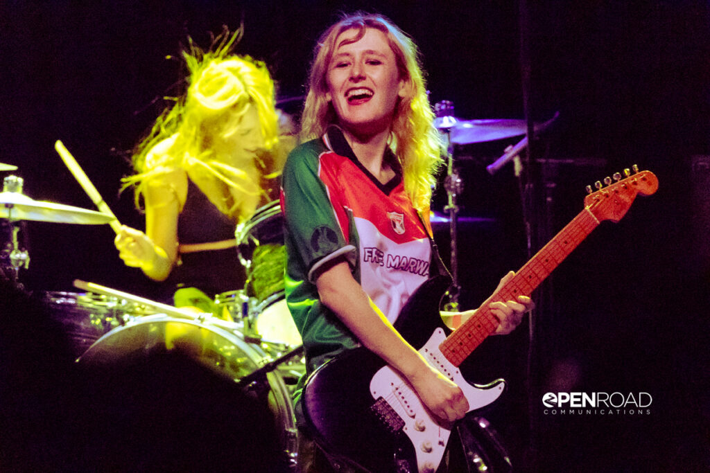 Lambrini Girls in concert. Singer Phoebe Lunny smiling holding a guitar on stage.
